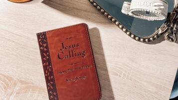 A wooden table with a brown leather journal titled "Jesus Calling," a dark brown mug, and a decorative mirror with a floral vase.