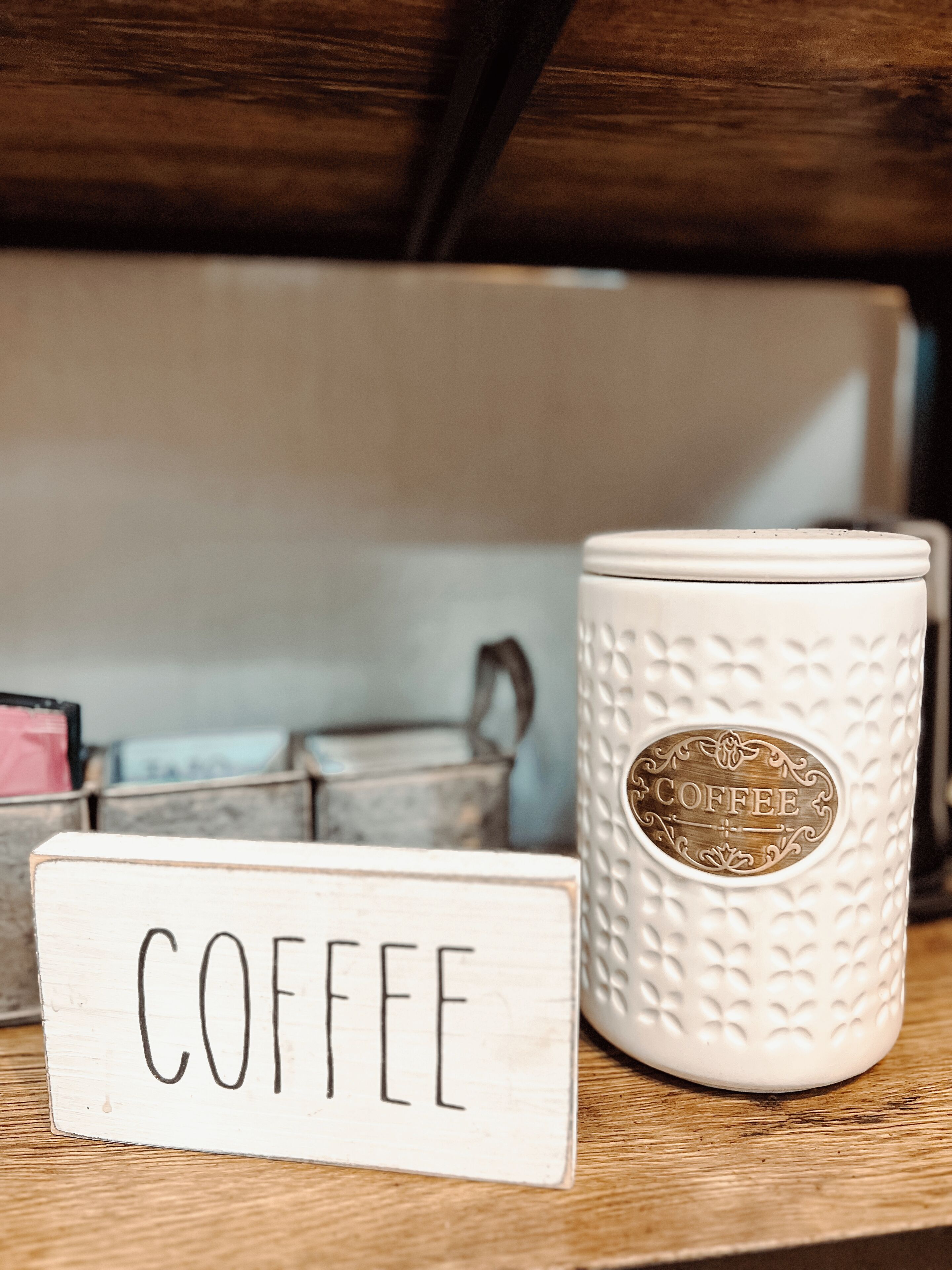 Une tasse en céramique blanche avec l’inscription « CAFÉ », une enseigne en bois portant le même mot, et un récipient en métal en arrière-plan.