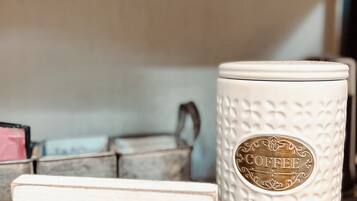 Une tasse en céramique blanche avec l’inscription « CAFÉ », une enseigne en bois portant le même mot, et un récipient en métal en arrière-plan.