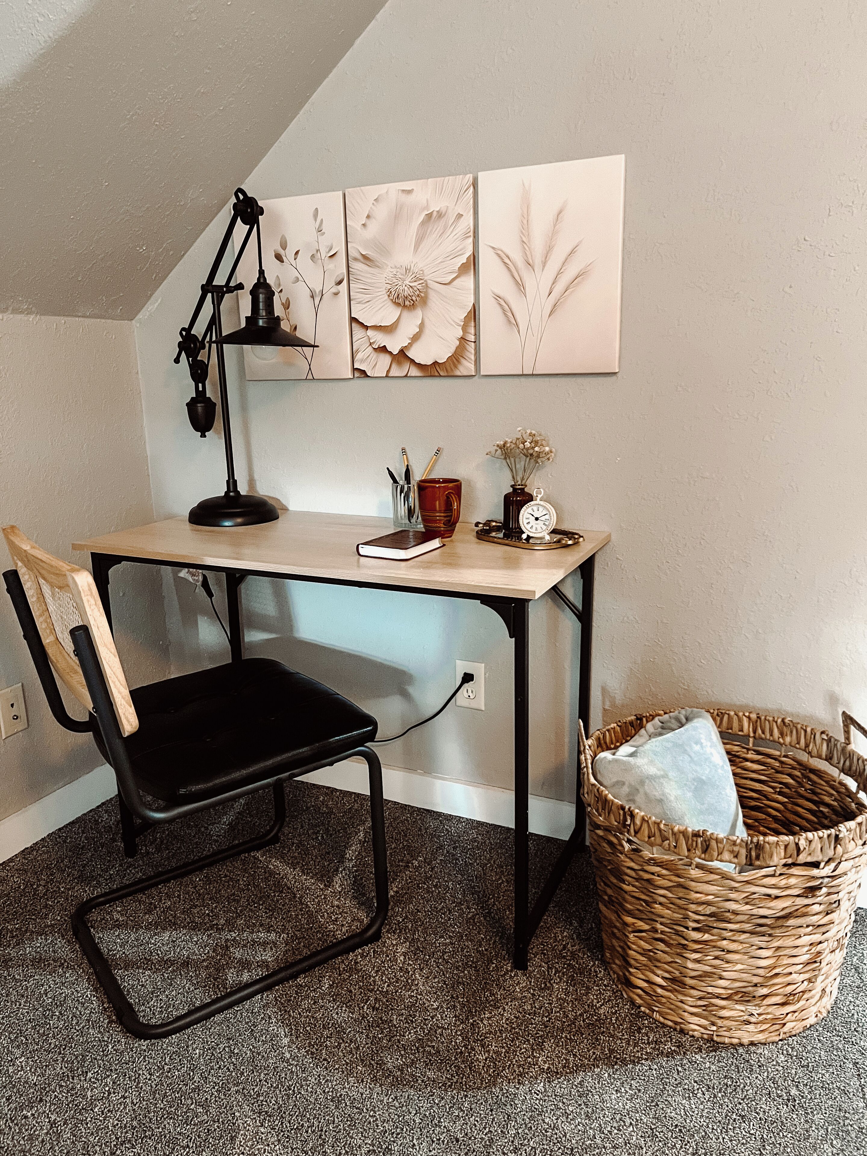 A corner desk with a black chair, a lamp, a book, a mug, a vase with flowers, and a wall with floral artwork.