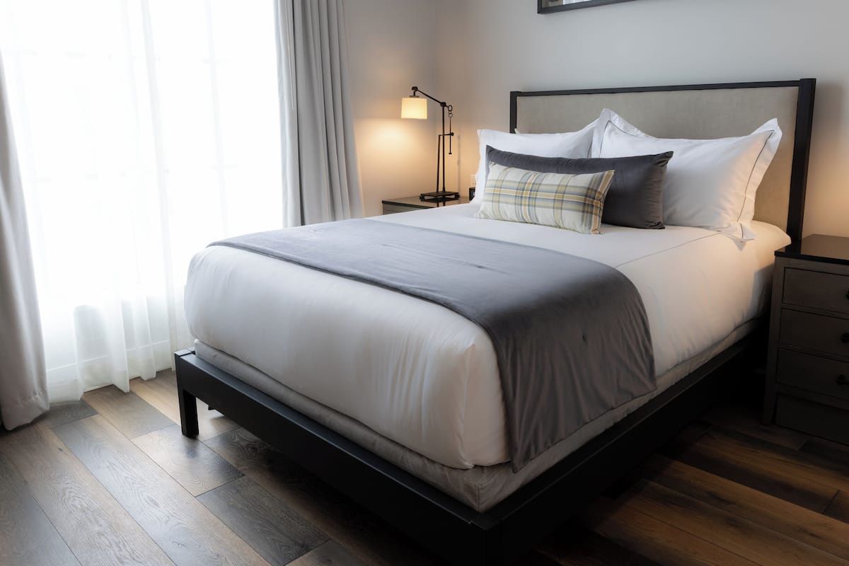 Una cama bien tendida con edredón gris y blanco, una lámpara de noche y una mesita de noche con cajón.