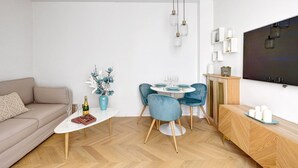 A modern living room with a sofa, a coffee table with a bottle and flowers, a dining area with a table and chairs, and a TV mounted on the wall. - 606 Aaron Suite - Apartment near the Eiffel Tower (Paris)