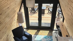 A modern interior with a wooden sloped ceiling, a black chair, a kitchen area with a sink, and a view of an outdoor deck with chairs and a grill. - Cabin 4: Reconnect with each other and nature at The Wildflower. (Princeton)
