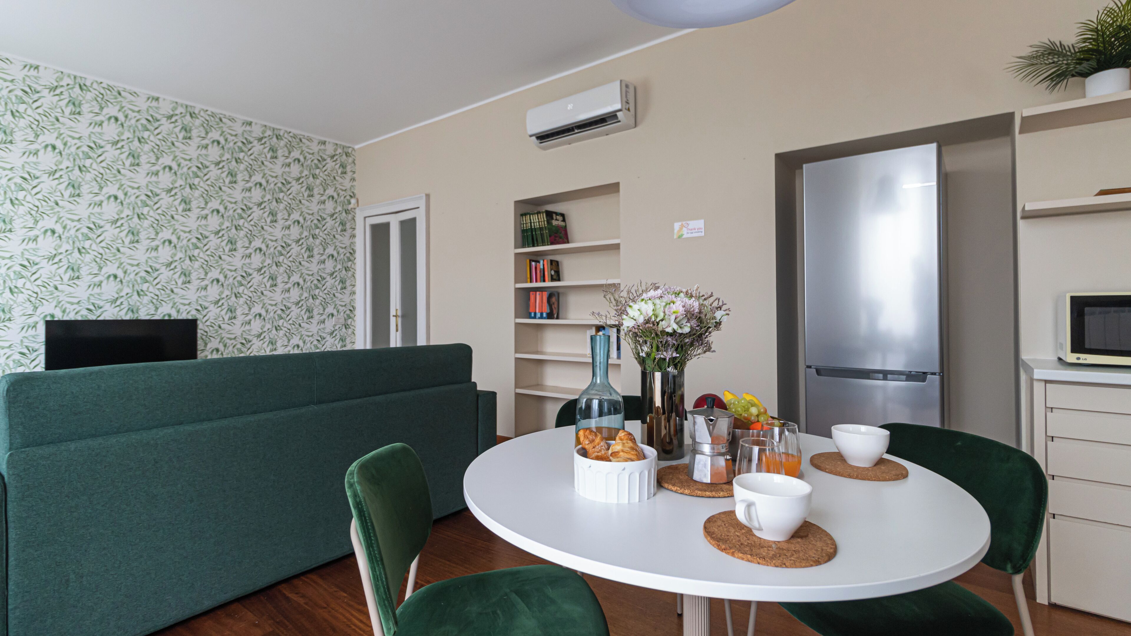 A modern living room with a green sofa, a white round dining table with chairs, a bookshelf, and a kitchen area with a refrigerator and microwave.