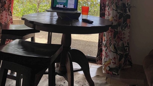 Une pièce avec un ordinateur portable posé sur une table, une télécommande et une tasse. On y trouve des rideaux à motifs floraux et une vue sur les arbres et le ciel.