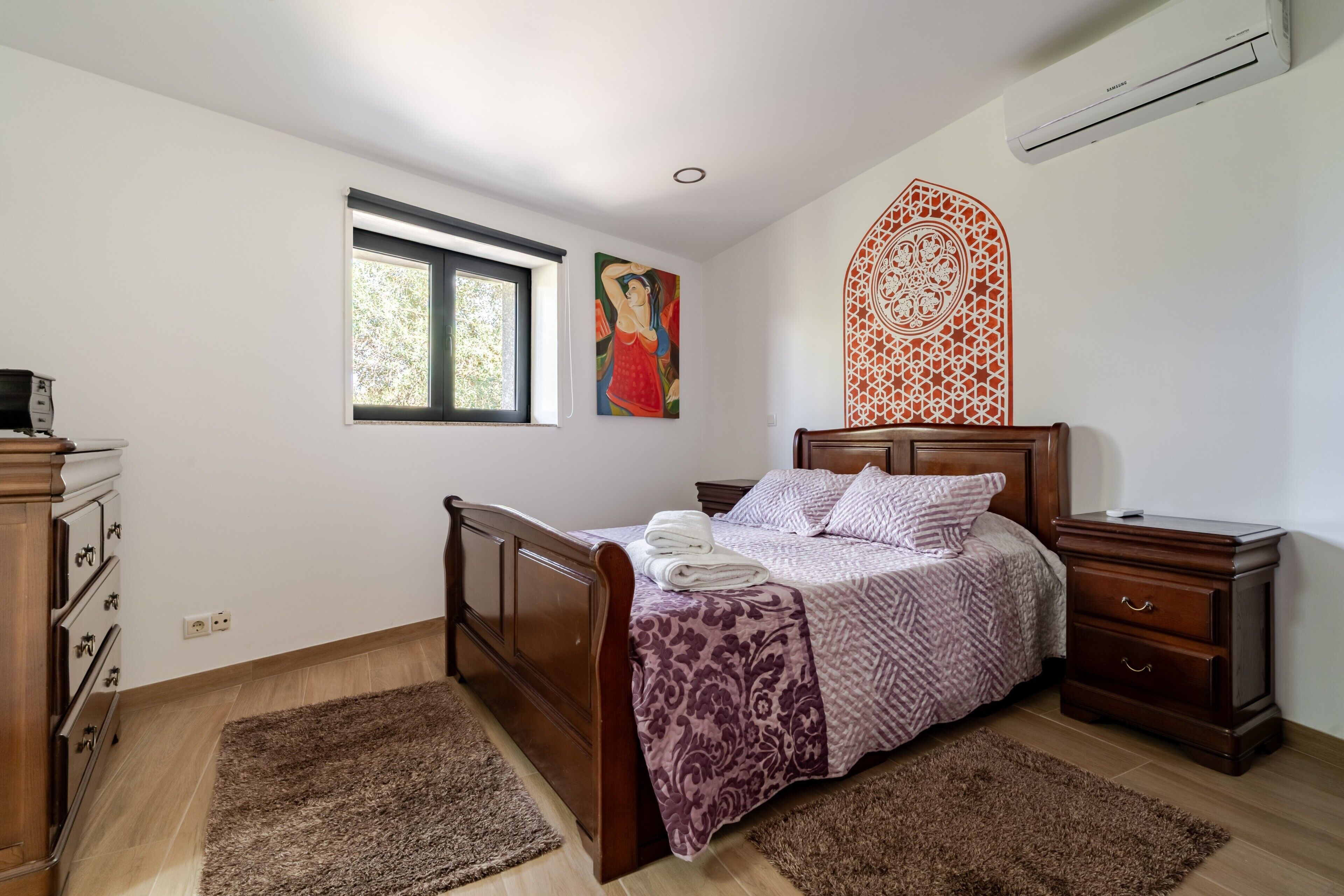 A bedroom with a wooden bed, a nightstand, a dresser, a window, and a wall decoration.