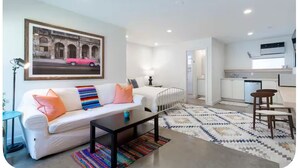 A modern apartment with a living area featuring a white sofa, colorful pillows, a coffee table, and a patterned rug. A kitchen with white cabinets and a dining area with a wooden table and chairs is visible. - Art-filled Cuban Studio 4 Creative Artsy Walkable Montrose Area. (Houston)