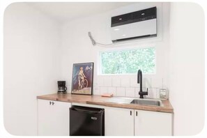 Une cuisine compacte avec des meubles blancs, un lave-vaisselle noir, une fenêtre donnant sur un espace verdoyant, un robinet noir et un appareil de climatisation.