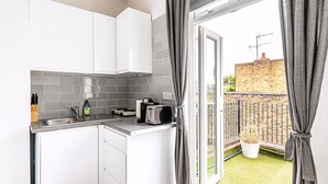 A compact kitchen with white cabinets, a sink, and a toaster. A window with curtains opens to a balcony with a brick wall and a potted plant. - North End Hideaway (London)