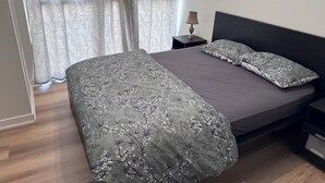 A bedroom with a large bed, two pillows, a bedside table with a lamp, and sheer curtains.