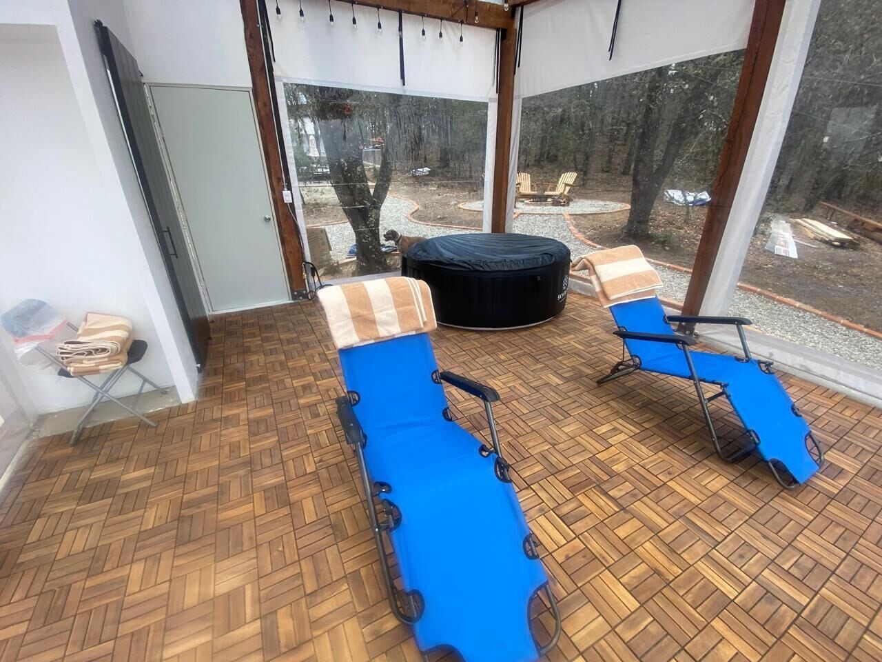 A sunroom with two blue lounge chairs, a wooden floor, and a view of a forested area outside.