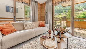 A modern living room with a beige sofa, a coffee table with drinks and snacks, and large windows offering a view of a forested area. - View village, balcony, tv, ski locker, 68m², Champagny-en-Vanoise (Champagny-en-Vanoise)