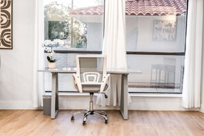Un espacio de oficina moderno con un escritorio, una silla y ventanas amplias que ofrecen vista a un edificio con techo de tejas.