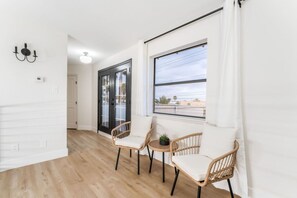 Ein Flur mit zwei Korbstühlen, einem kleinen Tisch und einer Topfpflanze. Eine Tür mit Glasfeldern und ein großer Fensterblick nach draußen.