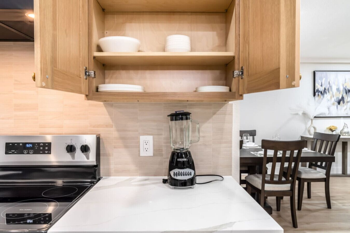 A modern kitchen with a built-in stove, a blender on the counter, wooden cabinets, and a dining area with chairs and a table.