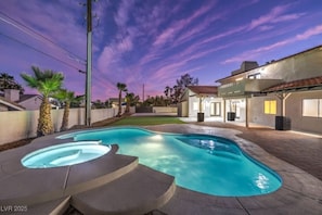 A swimming pool with a hot tub, surrounded by a patio area and a house with a red-tiled roof.