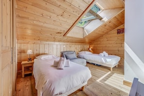 A cozy attic bedroom with two beds, wooden walls, a skylight, and a small table with a lamp.
