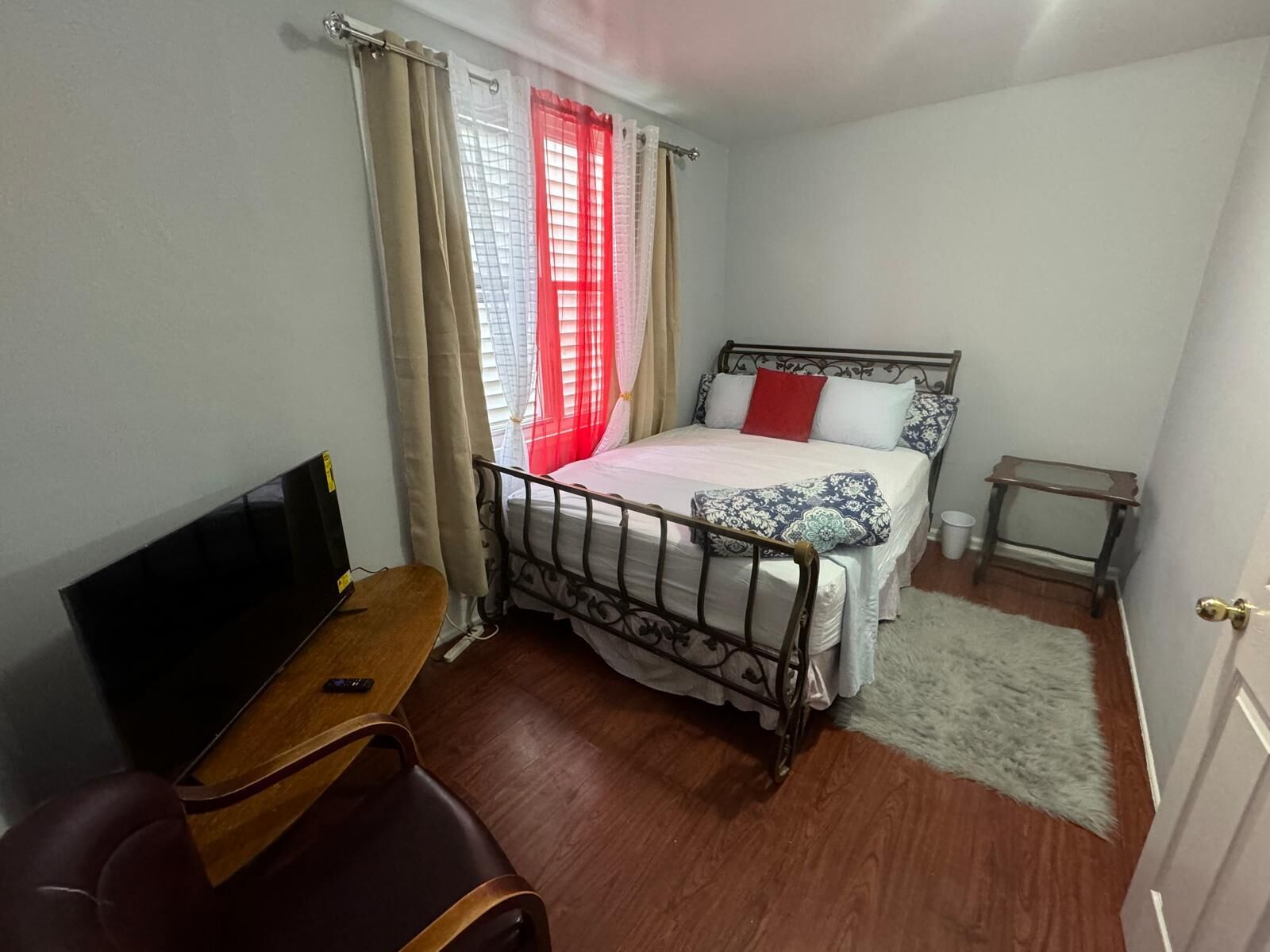 A bedroom with a bed, a TV, a chair, a small table, and a window with curtains.