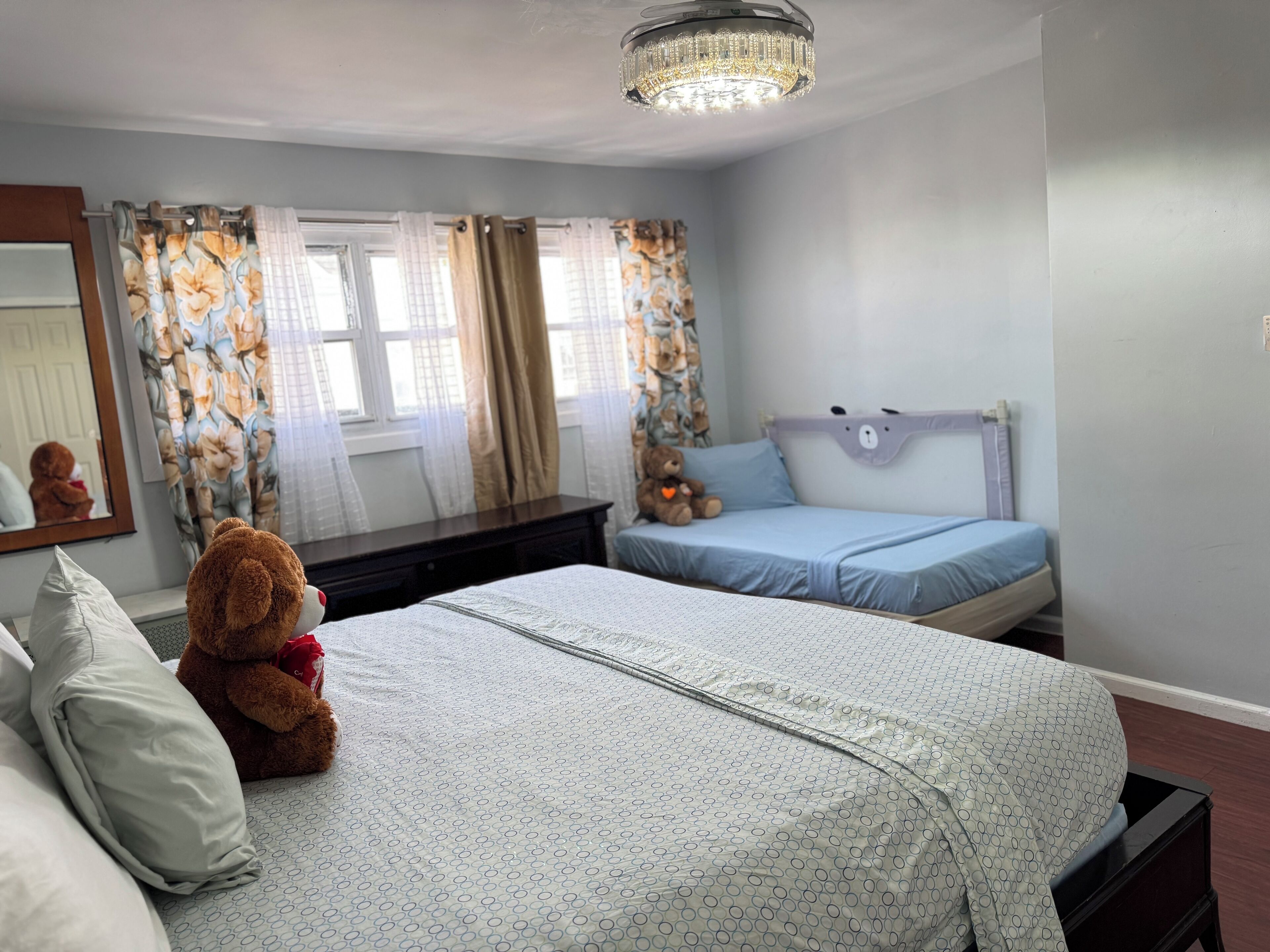 Ein Schlafzimmer mit Bett, Nachttisch, Schminktisch, Teddybär und einem Fenster mit Vorhängen.