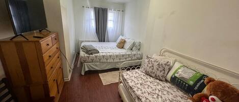 A small, well-lit room with two beds, a wooden dresser, a television, and a teddy bear.