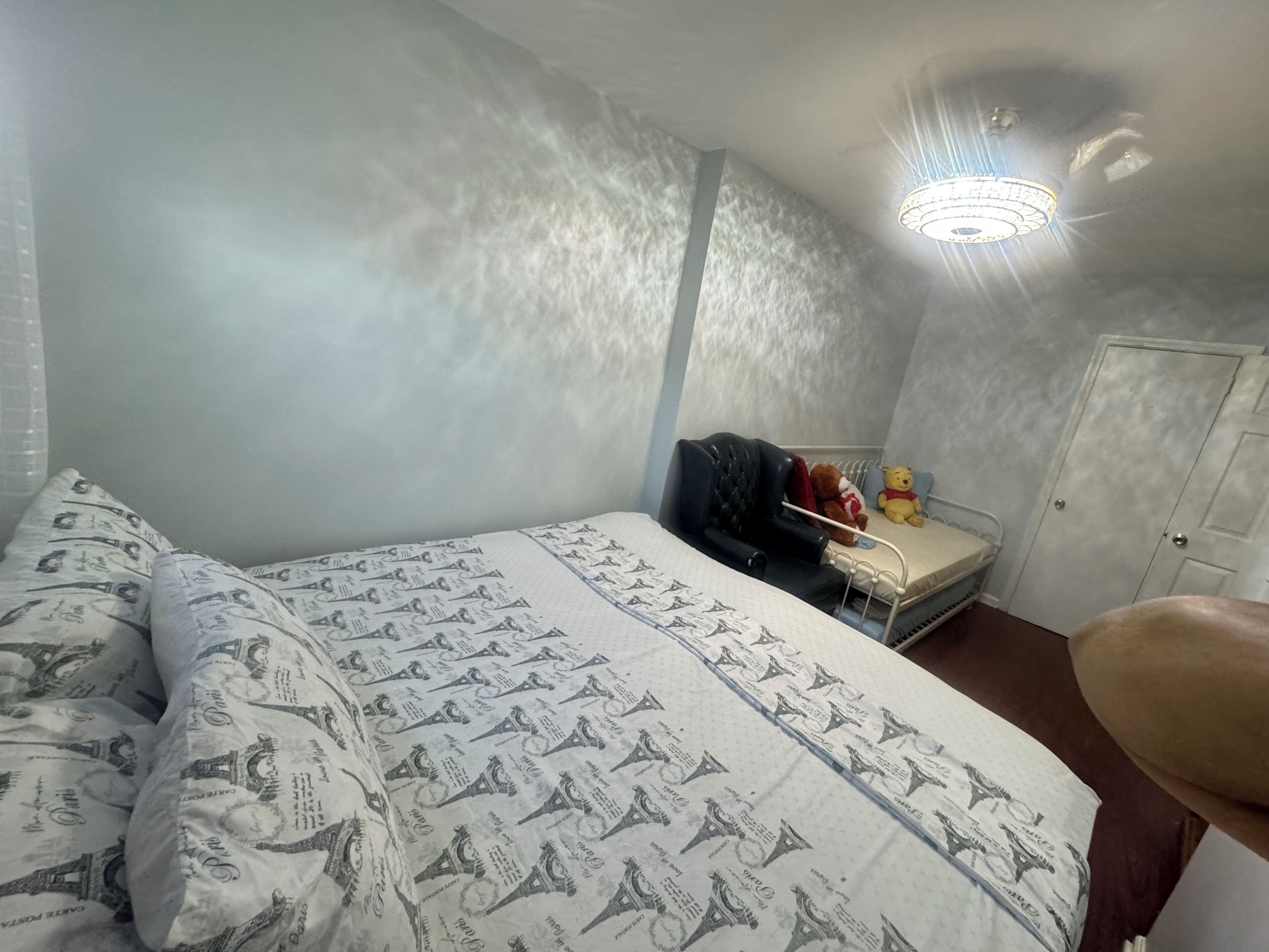 A bedroom with a bed, a nightstand, a chair, a teddy bear, and a door.