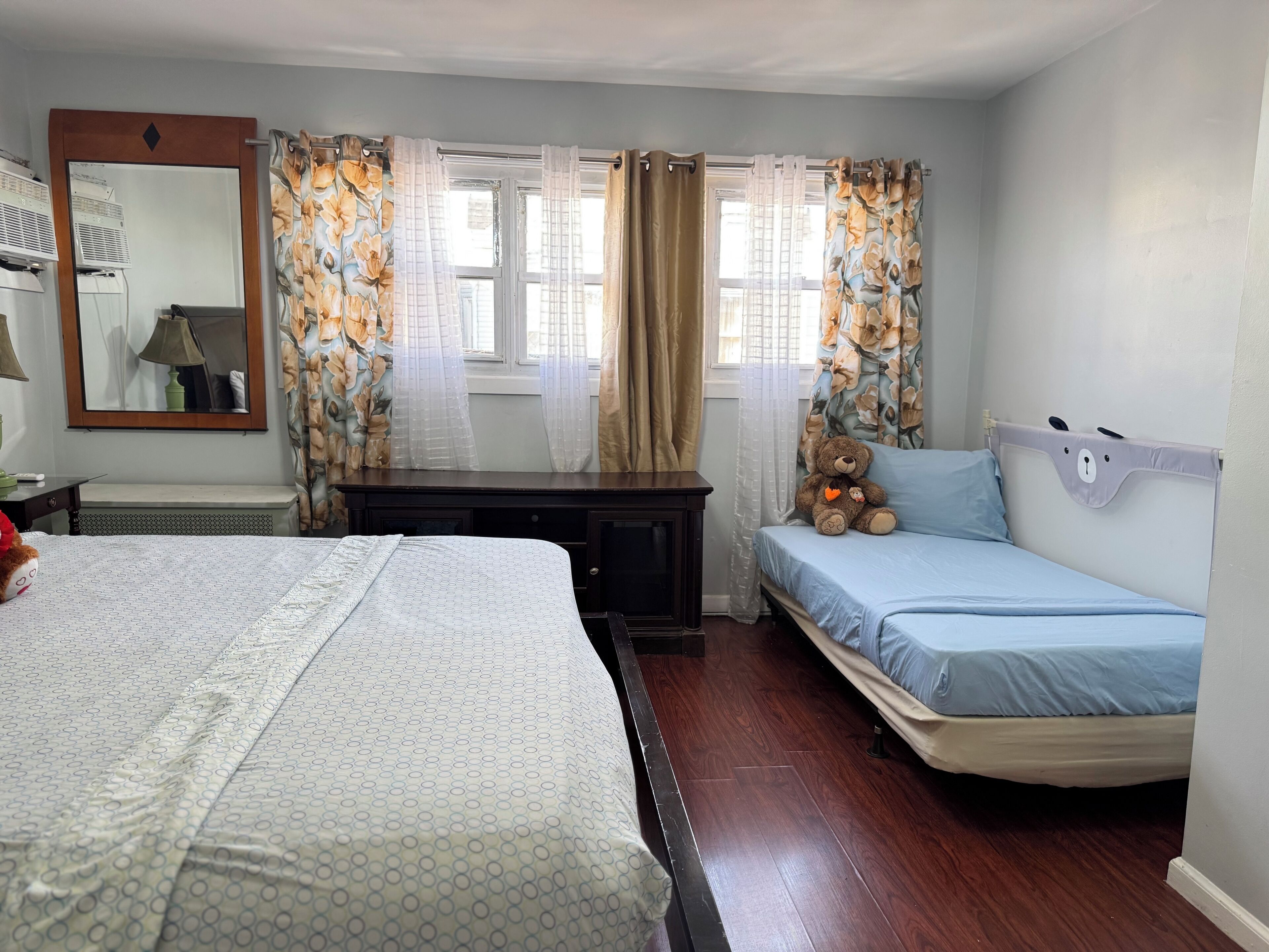 A bedroom with a bed, a small desk, a dresser, a window with curtains, and a teddy bear on a bed.