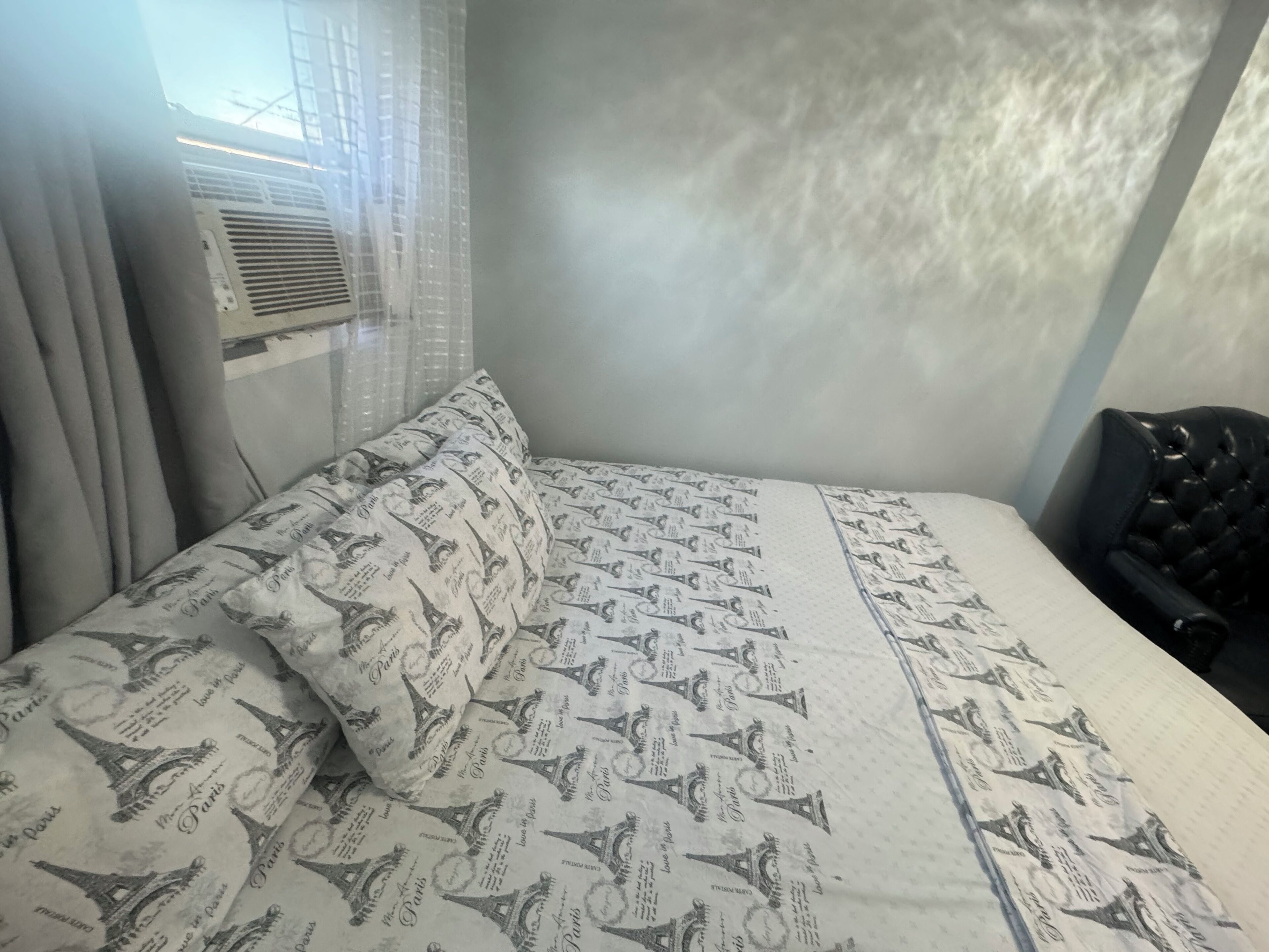 A bed with a patterned bedspread, a pillow, a black chair, and a window with curtains.