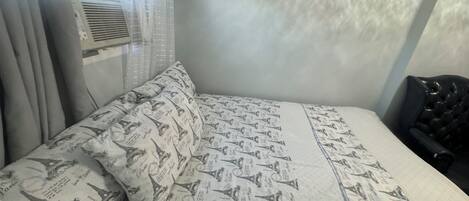 A bed with a patterned bedspread, a pillow, a black chair, and a window with curtains.
