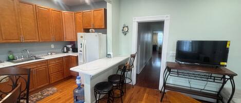 A compact kitchen with wooden cabinets, a white refrigerator, a sink, and a water dispenser. There is a dining area with a bench and chairs, and a TV on a stand.