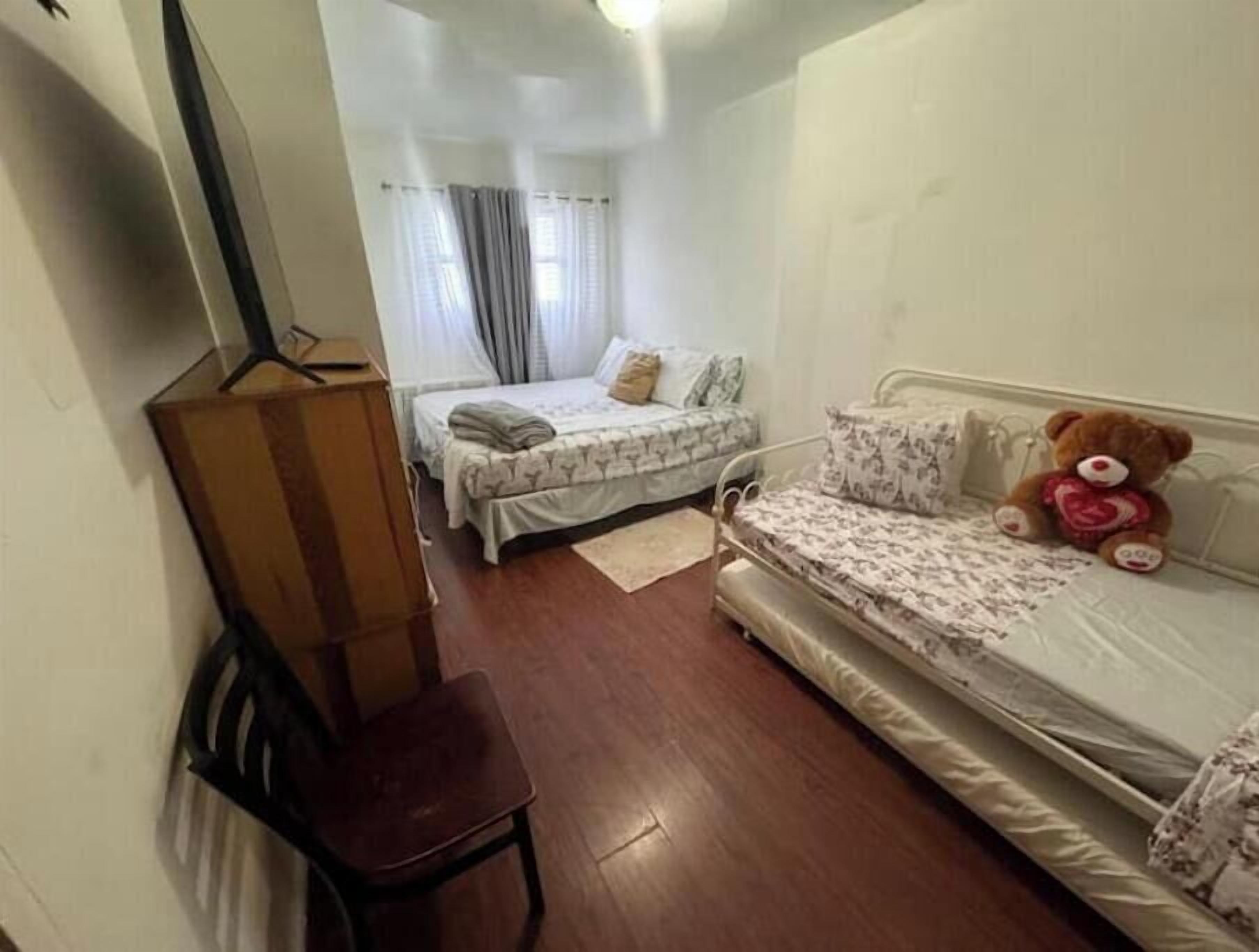 A small, compact room with two beds, a wooden chair, and a teddy bear.