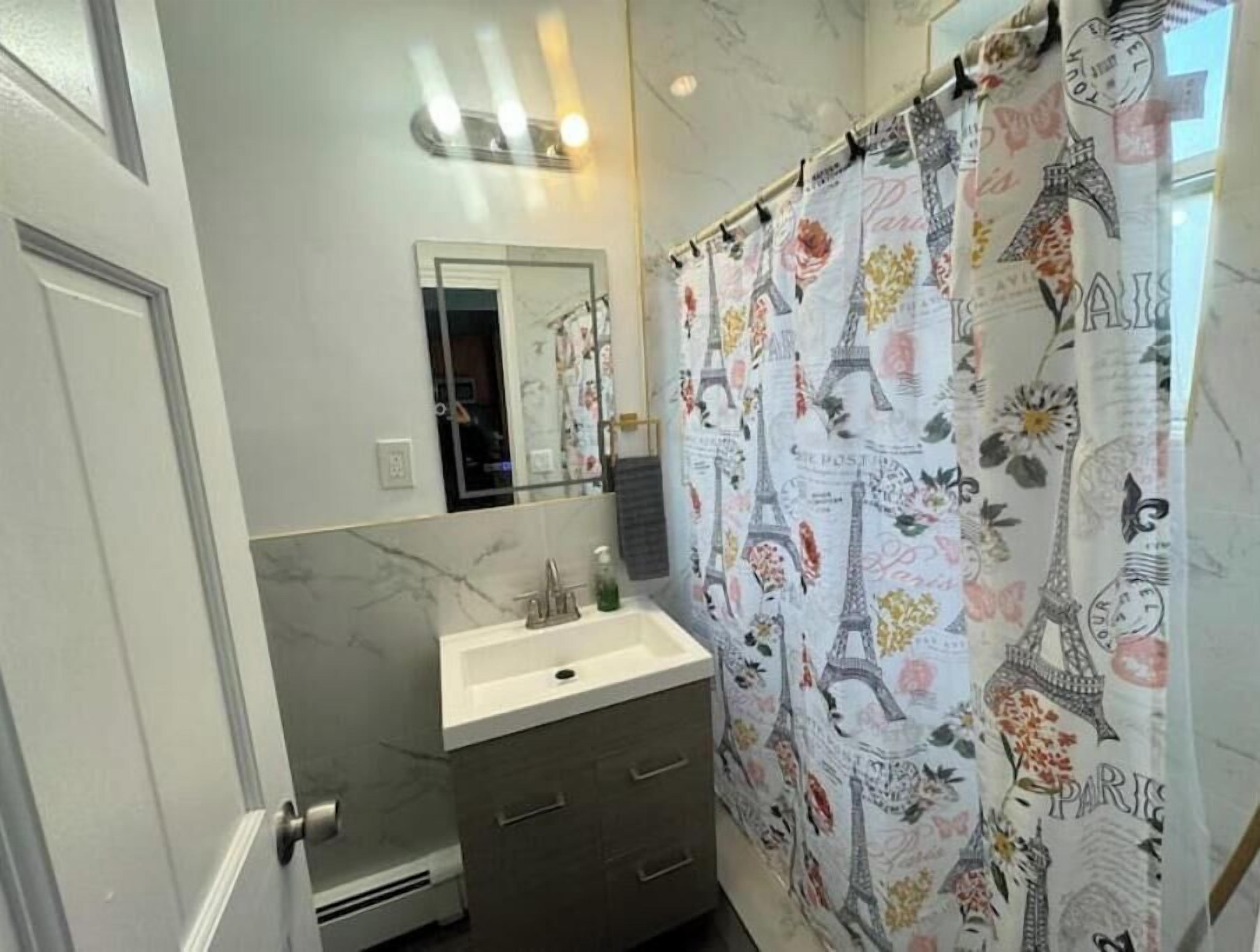 A bathroom with a marble wall, a sink with a mirror, and a shower curtain with Paris-themed designs.