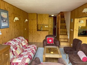 Un interior de madera con un sofá, sillones, una mesa de centro y una escalera que lleva a una zona de cocina.