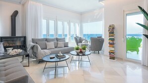 A modern living room with a sofa, two armchairs, a coffee table, and a fireplace. There is a large window with a view of the sea.