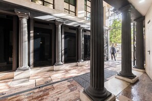 Un espacio exterior techado con suelo de mármol y columnas clásicas, que conduce a la entrada de un edificio.