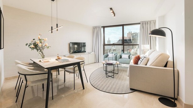 A modern living room with a dining area, a sofa, a TV, and a large window with a city view. - Constitución III - Two Bedroom Apartment, Sleeps 4 (Malaga)