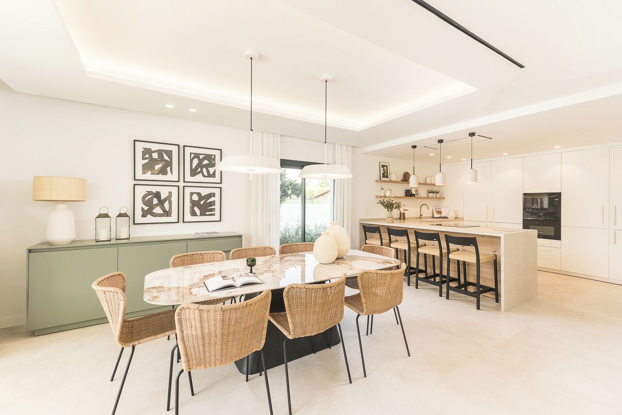 A modern dining area with a round table, wicker chairs, pendant lights, and a view of the kitchen.