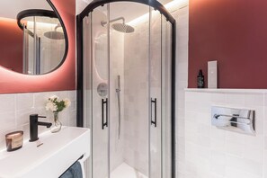 Ein modernes Badezimmer mit einer ein Glaskabine abgetrennten Dusche, einem weißen Waschbecken mit schwarzem Wasserhahn und einem Spiegel mit rundem Rahmen.