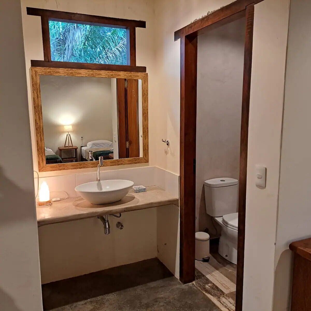 A bathroom with a large mirror, a white sink, a toilet, and a small trash bin.