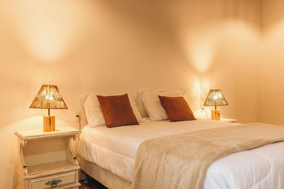 A neatly made bed with two lamps on either side, a white wall, and a small wooden nightstand with a drawer.