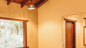 Une chambre avec un bureau en bois, une chaise, un lit recouvert d’une couverture blanche, une fenêtre donnant sur des arbres et une porte en bois.