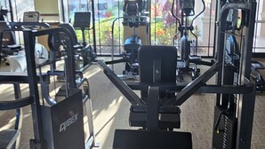 Ein Fitnessstudio mit verschiedenen Trainingsgeräten, großen Fenstern und einem Teppichboden.