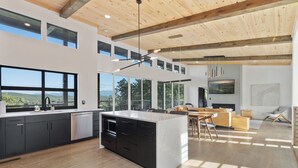 A modern kitchen with a large island, black cabinets, and a dishwasher. There is a dining area with a wooden table and chairs. The room has large windows and a view of the outdoors. - Smoky Vista / Hot Tub & Mountain Views (Sevierville)
