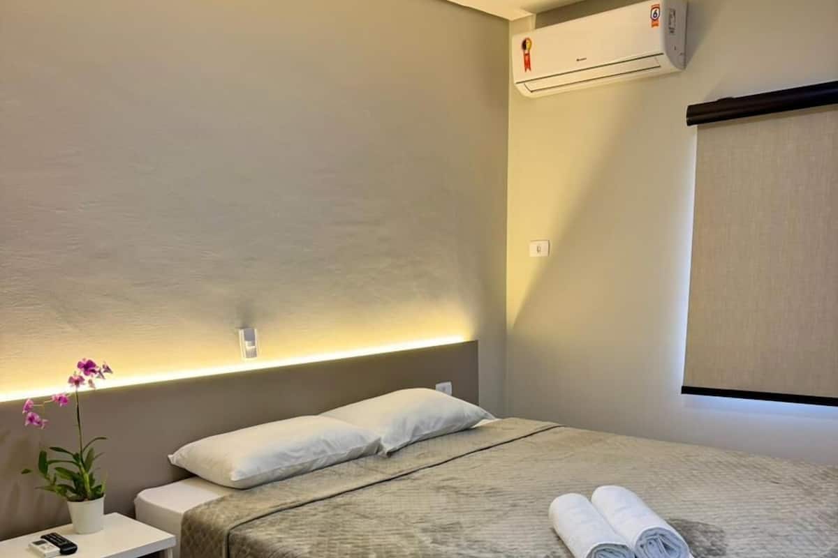 Una habitación de hotel ordenada con cama, mesita de noche, silla y equipo de aire acondicionado.