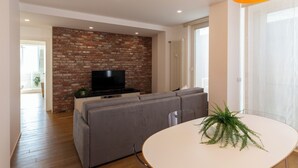 A modern living room with a brick accent wall, a grey sofa, a flat-screen TV, and a round dining table with a plant. - notaMI - HELLO Navigli (Milan)
