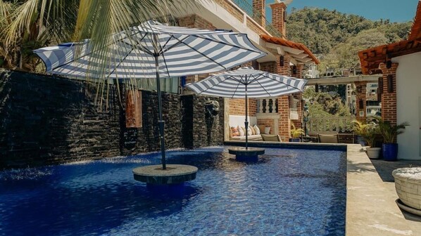 A swimming pool with two striped umbrellas, a stone wall, and a view of a hillside with buildings.