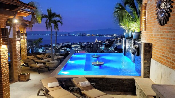 Una piscina infinita al aire libre con sillones de descanso, vista al paisaje urbano y un muro de ladrillo con detalles decorativos.