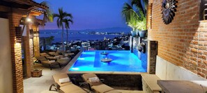 An outdoor infinity pool with lounge chairs, a view of the cityscape, and a brick wall with decorative elements.