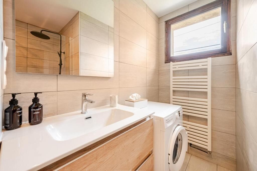 Un baño moderno con lavamanos blanco, armarios de madera, ducha integrada y ventana sobre el perchero.