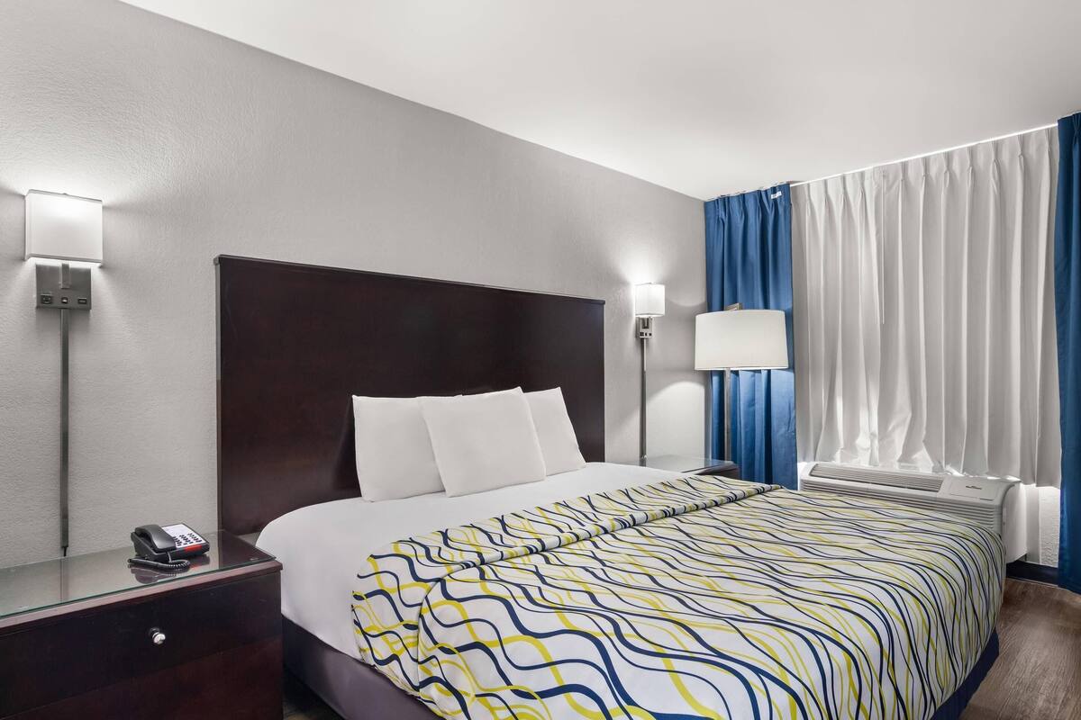 A hotel room with a large bed, a bedside table with a phone, a wall-mounted lamp, and a window with curtains.
