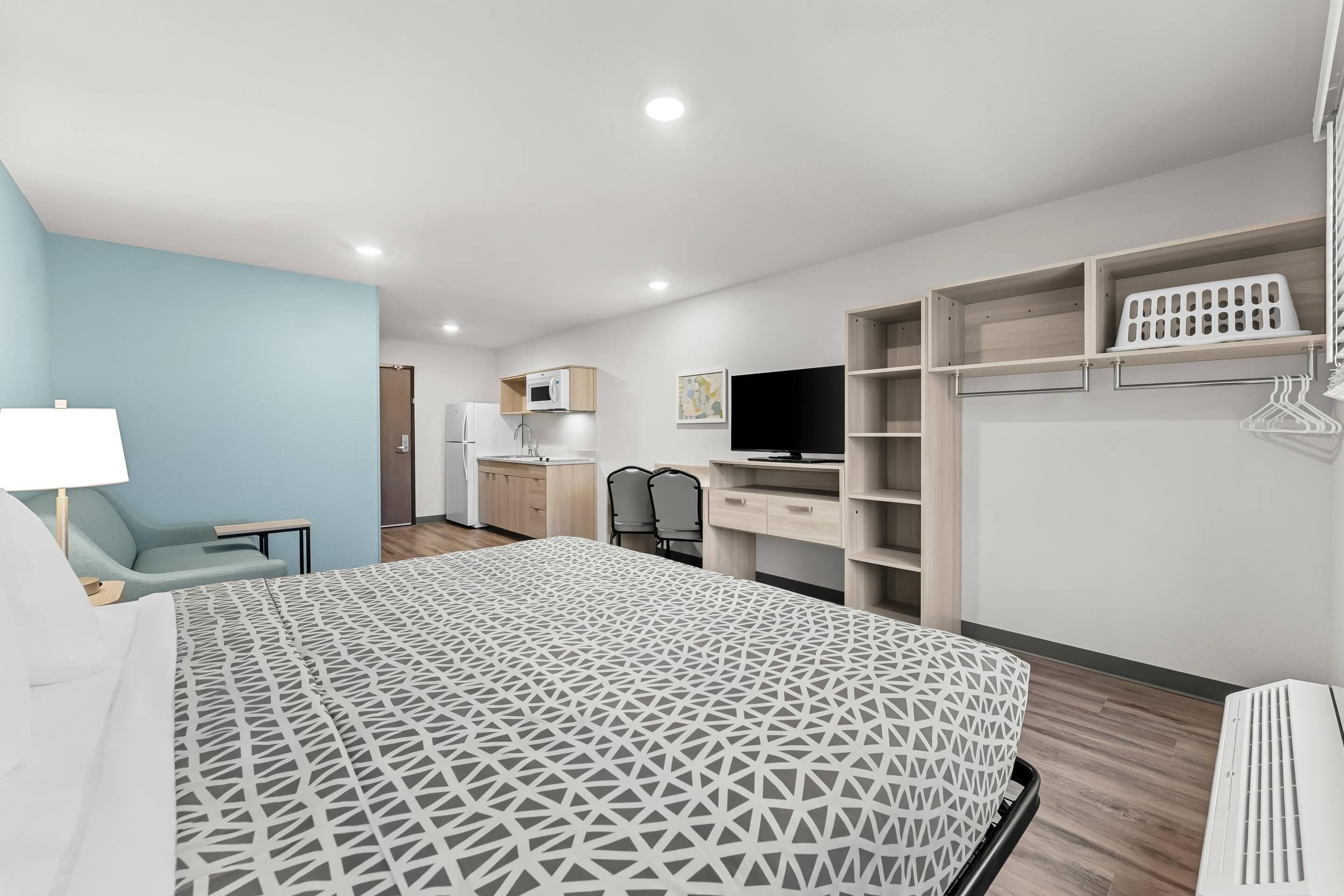 A hotel room with a bed, a desk, a chair, a TV, a wardrobe, and a kitchenette.